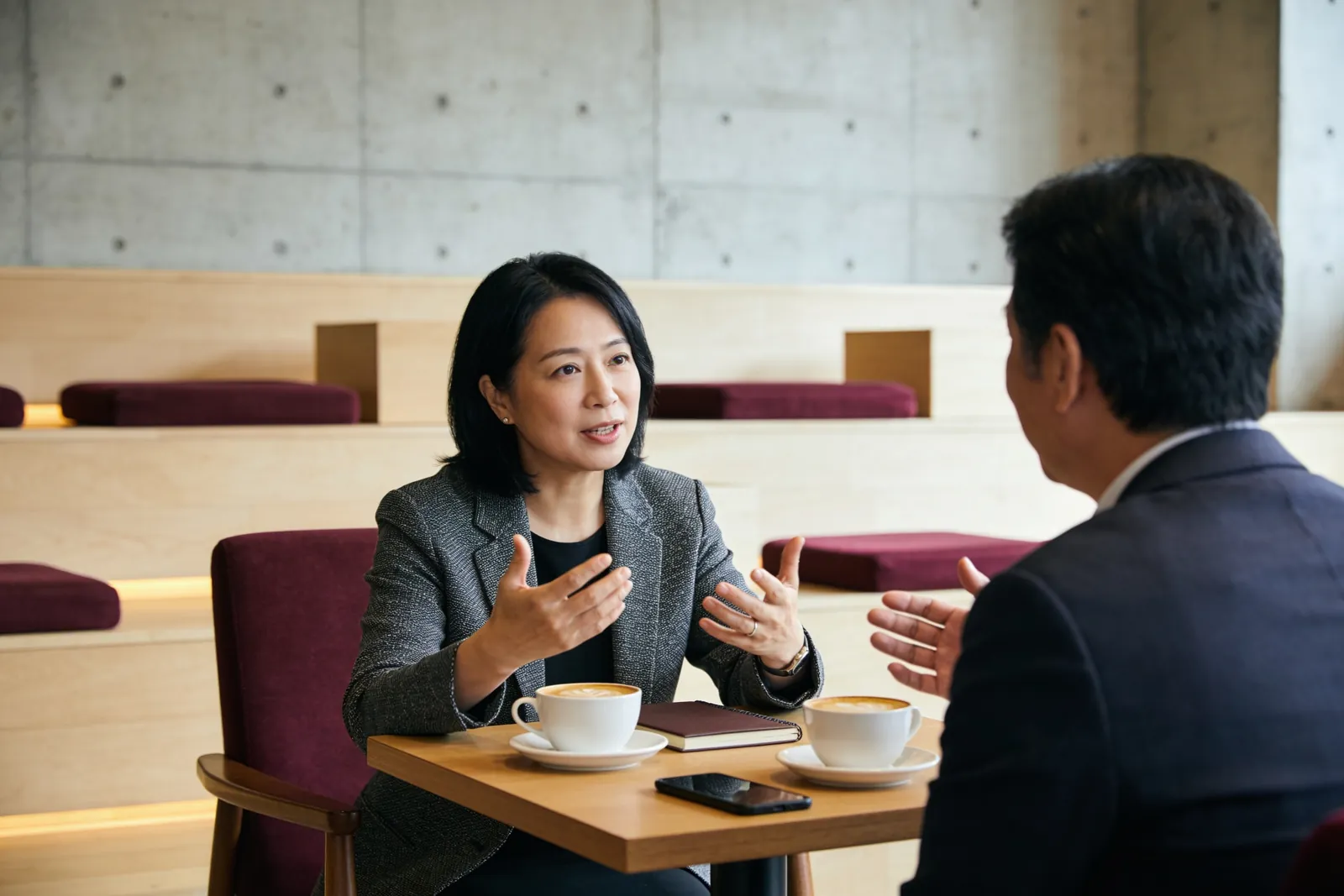咖啡館裡兩位成員面對面坐著,女士一邊比手勢一邊說明,男士在聽,桌上兩杯咖啡和一本筆記本。